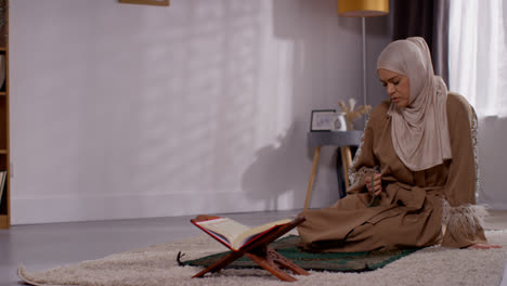 Mujer-Musulmana-Con-Hijab-Rezando-Con-Cuentas-De-Oración-En-Casa-Sobre-Una-Alfombra-De-Oración-Con-Una-Copia-Abierta-Del-Corán-En-El-Stand